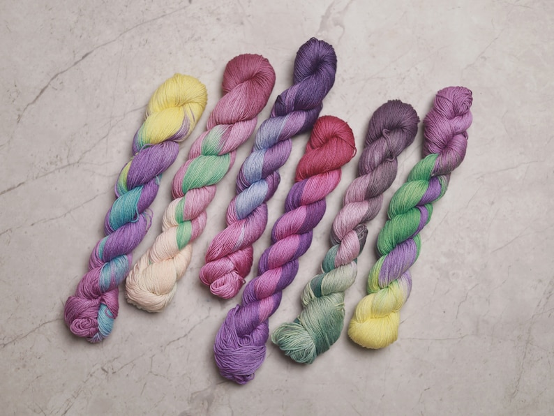 May include: Seven skeins of yarn in various shades of purple, pink, green, and yellow. The yarn is twisted and arranged in a fan shape.