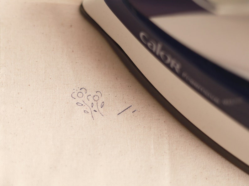 May include: A white iron with the word "Calor" printed on it is being used to press a small floral design onto a piece of white fabric.