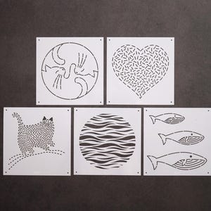 Puede incluir: Un conjunto de cinco plantillas cuadradas blancas con varios diseños recortados. Los diseños incluyen dos gatos formando un círculo, un corazón relleno de rayas, un gato con pelaje texturizado, un patrón circular de líneas onduladas y tres ballenas.