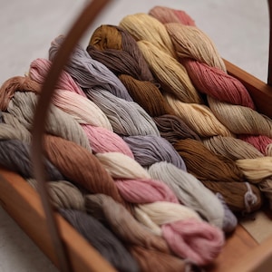 May include: A collection of natural colored yarn skeins in a wooden basket. The yarn is arranged in a rainbow pattern, with shades of brown, beige, pink, and gray.
