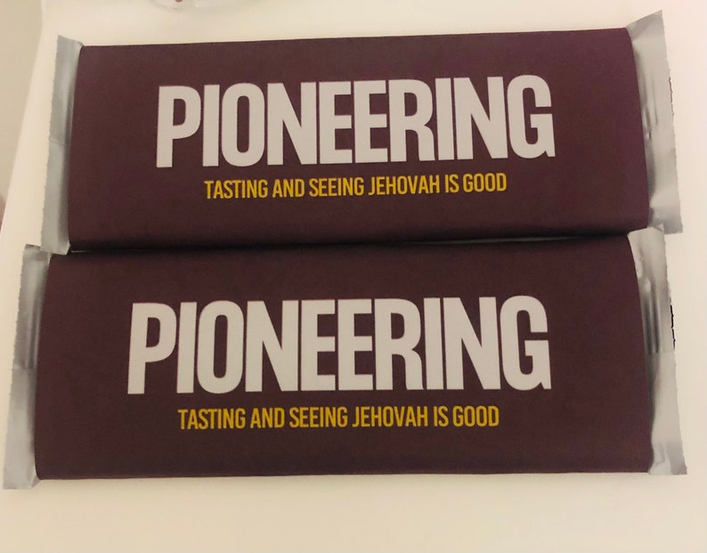 May include: Two chocolate bars in dark brown packaging with the word "PIONEERING" in large white letters. Below, the text reads "TASTING AND SEEING JEHOVAH IS GOOD" in a smaller, gold font. The bars are wrapped in silver foil.