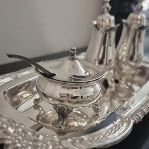 May include: A silver-plated serving set, comprising a lidded bowl with a spoon, two shakers, and a decorative tray. The set features ornate detailing and is presented on a dark surface. The pieces reflect the surrounding environment.