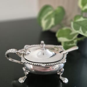 May include: A small, ornate silver serving dish with a lid and a small spoon. The dish has a handle and sits on three decorative feet. The rim of the dish and the lid are detailed with a scalloped design. The spoon rests on the lid.