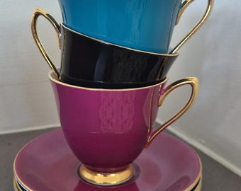 May include: Three stacked teacups with gold trim. The top cup is teal, the middle cup is black, and the bottom cup is a deep purple. The cups are sitting on a matching saucer.