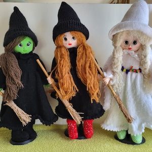 Op de afbeelding: Drie handgemaakte heks poppen met zwarte hoeden en jurken. Eén pop heeft een groene huid en bruin haar, de tweede heeft rood haar en rode schoenen, en de derde is wit met blond haar. Elke pop houdt een bezem vast.