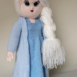 Pode incluir: Uma boneca de tricô com cabelo branco comprido, vestindo um vestido azul claro e um casaco combinando. A boneca tem olhos azuis e lábios vermelhos.
