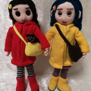 Könnte beinhalten: Zwei Strickpuppen, eine in einem roten Mantel mit einer schwarzen Katze in einer gelben Tasche und die andere in einem gelben Regenmantel mit einer braunen Tasche.