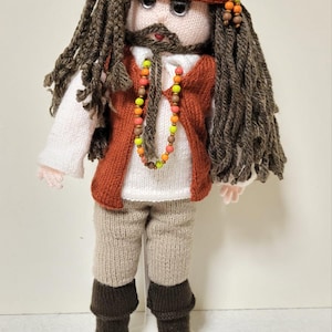 Puede incluir: Una muñeca de punto con cabello castaño, un gorro rojo, una camisa blanca, un chaleco rojo, pantalones marrones y botas marrones. La muñeca lleva un collar de cuentas de colores.
