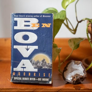 May include: A blue paperback book with white lettering that reads "BEN BOVA MOONRISE" and "SPECIAL REBATE OFFER - SEE INSIDE". The book is sitting on a wooden table next to a potted plant and a rock.