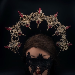 May include: A gold-colored crown with red crystal accents and spikes. The crown is ornate with a filigree design and is set against a black background. The crown is a gothic or fantasy-themed accessory.