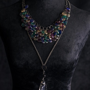 Può includere: Collana multistrato esposta su un manichino nero. Lo strato superiore presenta un design a pettorina con elementi circolari iridescenti nelle tonalità del blu, del verde e del viola. Una catena più lunga con un pendente trasparente a forma di goccia pende sotto.