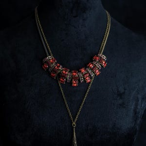 May include: A vintage-style necklace with a dark bronze chain and a series of red rectangular and square-cut gemstones. The necklace has a pendant with a red crystal. The jewelry is displayed on a black mannequin.