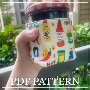 Peut inclure: Un bocal avec un couvercle à carreaux rouges et blancs et un corps noir, enveloppé dans une housse en tissu. La housse présente un motif répété de piments, de bouteilles de sauce piquante et le mot "HOT". Le texte "PDF PATTERN" est en bas.