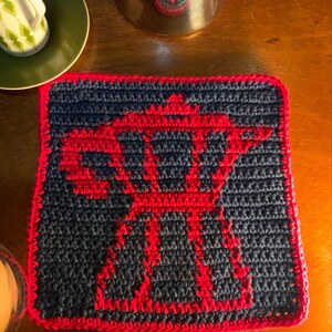 Op de afbeelding: Handgemaakte gehaakte onderzetter met een rood en marineblauw ontwerp. De onderzetter heeft een rode rand en een centraal patroon. Deze vierkante onderzetter is ideaal om oppervlakken te beschermen tegen warme of koude dranken.