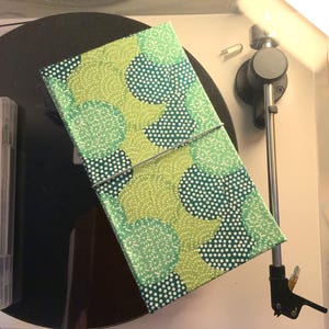Può includere: Un diario a fantasia con un design verde e bianco, fissato con una chiusura a filo, poggia su un giradischi. La copertina del diario presenta un motivo ripetuto di cerchi e punti nelle tonalità del verde e del bianco.