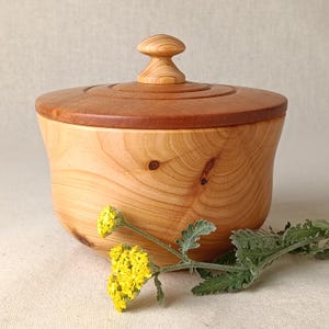 May include: A wooden lidded bowl, featuring a light-coloured body and a darker, reddish-brown lid. The lid has a small, rounded knob. The bowl is set against a neutral background, with yellow flowers and green foliage in the foreground.