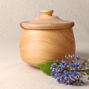 May include: A light-coloured wooden jar with a lid. The jar has a rounded body and a lid with a small knob. A sprig of blue flowers and a green leaf are in the foreground. The wood grain is visible.