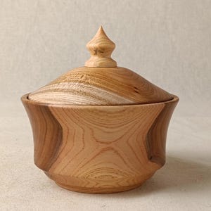 May include: A small, wooden box with a lid. The box is made of light-coloured wood with a natural finish. The lid has a pointed knob on top. The box is a decorative item.