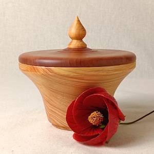 May include: A wooden lidded container with a light brown body and a darker brown lid. The lid is topped with a decorative finial. A vibrant red flower with a yellow centre rests beside the container, adding a pop of colour.