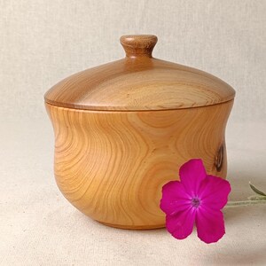 May include: A wooden bowl with a lid, crafted from light-coloured wood. The natural grain is visible on the bowl and lid. A small, round knob acts as a handle. A vibrant pink flower is positioned in front of the bowl.