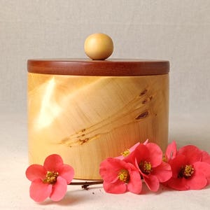 May include: A cylindrical wooden container with a light-coloured, polished grain body and a darker, flat circular lid topped with a small, spherical wooden knob. Several bright pink flowers with yellow centres are positioned in front of the base.