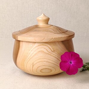 May include: A wooden jar with a lid, showing a light wood grain. The jar has a rounded shape and a small, pointed knob on the lid. A bright pink flower is placed beside the jar, adding a splash of colour to the scene.