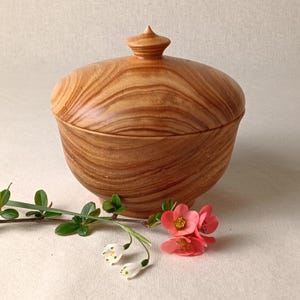 May include: A wooden bowl with a lid, showing a natural wood grain pattern. The bowl is light brown with a rounded shape and a small, pointed knob on the lid. Greenery and pink flowers are in the foreground.