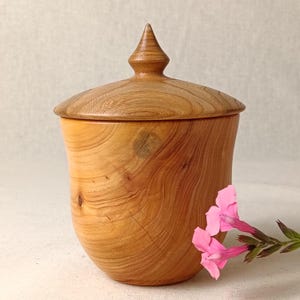 May include: A wooden jar with a lid and a pointed top. The jar is made of light-coloured wood, showing natural grain patterns. A sprig of pink flowers is placed beside the jar, adding a splash of colour.