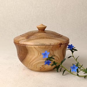 May include: A wooden bowl with a lid, displaying intricate wood grain. The bowl has a curved form and a small knob on the lid. A sprig of blue flowers adds a splash of colour. The bowl is likely made from a hardwood.