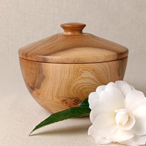 May include: A wooden bowl with a lid, made from light-coloured wood. The bowl has a rounded shape with a lid featuring a small knob. A white flower and green leaf are in the foreground. The bowl is likely for storage or decorative purposes.