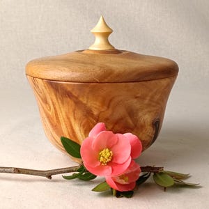May include: A wooden bowl with a lid, showcasing a light-coloured finial. The bowl has a natural wood grain pattern. Pink flowers and green leaves are arranged nearby, creating a decorative display.