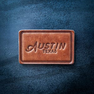 May include: A rectangular, brown leather patch with the city name "Austin" and the state "Texas" embossed in a cursive font. The patch has a darker brown border and is set against a dark blue textured background.