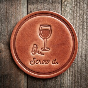 May include: A round, brown leather coaster with a debossed wine glass and bottle opener design. The phrase "Screw it." is also debossed on the coaster. The coaster is set against a wooden background.