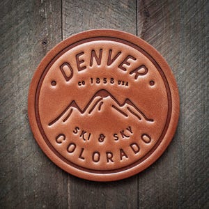 May include: A round, brown leather coaster with the embossed text "DENVER CO 1858 USA" and "SKI & SKY COLORADO" surrounding a mountain range graphic. The coaster is circular and has a detailed, textured surface.