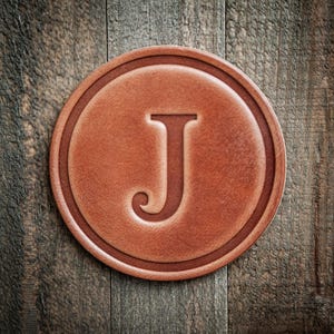 May include: A round, brown leather coaster with the letter "J" embossed in the center. The coaster has a raised border and is set against a rustic wooden background. The color is a warm, reddish-brown.