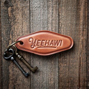 May include: A brown leather keychain tag with the word "YEEHAW!" embossed on it. The tag is attached to a metal ring with two antique-style keys. The background is a rustic wooden surface.