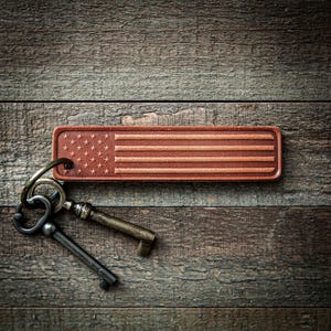 May include: A brown leather keychain with an embossed American flag design. The keychain is attached to a set of antique keys. The background is a rustic wooden surface.