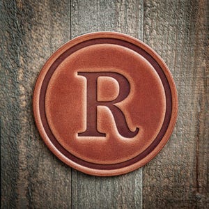 May include: A round, brown leather coaster with a raised border and a large letter "R" in the center. The coaster is set against a rustic wooden background, creating a warm, inviting aesthetic.