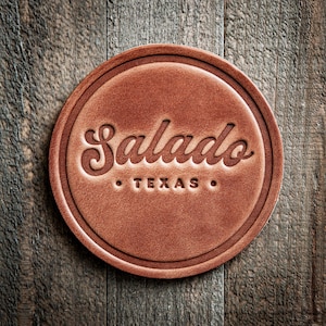 May include: A round, brown leather coaster with the word "Salado" in a decorative script, and "TEXAS" below it. The coaster is set against a rustic wooden background.