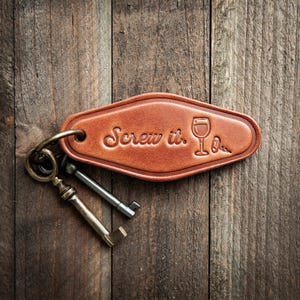 May include: A brown leather keychain with the phrase "Screw it." and a wine glass illustration. The keychain is attached to three keys and rests on a wooden surface.