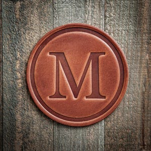 May include: A round, brown leather coaster with a debossed letter "M" in the center. The coaster has a raised border and sits on a weathered wooden surface. The leather has a rich, textured appearance.