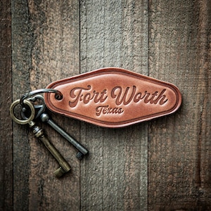 May include: A brown leather key tag with the words "Fort Worth Texas" embossed on it. The tag is attached to a metal keyring with two antique keys. The background is a rustic wooden surface.