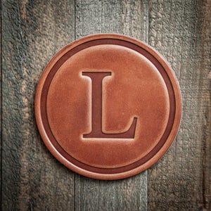 May include: A round, brown leather coaster with the letter "L" embossed in the center. The coaster has a raised border and sits on a weathered wooden surface.