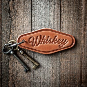 May include: A brown leather keychain with the word "Whiskey" embossed on it, attached to two antique keys. The keychain is oval-shaped and hangs against a rustic wooden background.
