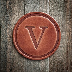 May include: A round, brown leather coaster with a debossed letter "V" in the center. The coaster has a raised border and is set against a rustic wooden background. The leather has a textured appearance.