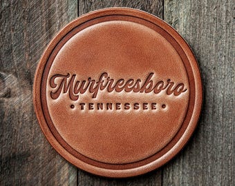 May include: A round, brown leather coaster with the embossed text "Murfreesboro Tennessee". The coaster has a decorative border and is set against a rustic wooden background.
