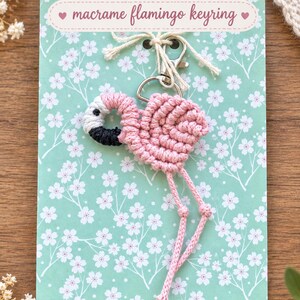 May include: A macrame flamingo keyring featuring pink cord knotted into a flamingo shape with a white and black head, attached to a silver metal lobster clasp. The keyring is displayed on a light green card with white floral patterns and the text macrame flamingo keyring.