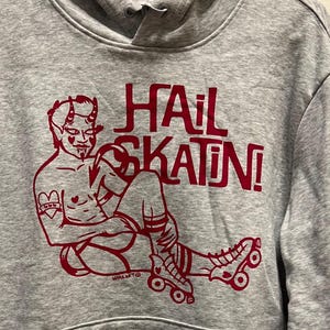 Puede incluir: Una sudadera gris con un gráfico rojo de una figura diabólica sobre patines, con las palabras "HAIL SKATIN!" en rojo. La sudadera tiene un bolsillo delantero y una capucha con cordón.