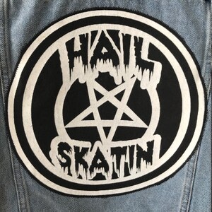 May include: A black and white patch with the words "HAIL SKATIN" in a dripping font, surrounding a white pentagram. The patch is circular and set against a light blue denim jacket.