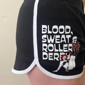 Könnte beinhalten: Schwarze Sporthosen mit weißem Besatz und einem weißen Streifen an der Seite. Die Shorts zeigen den Text "BLOOD, SWEAT & ROLLER DERBY" mit einer Grafik eines Roller-Derby-Mädchens.